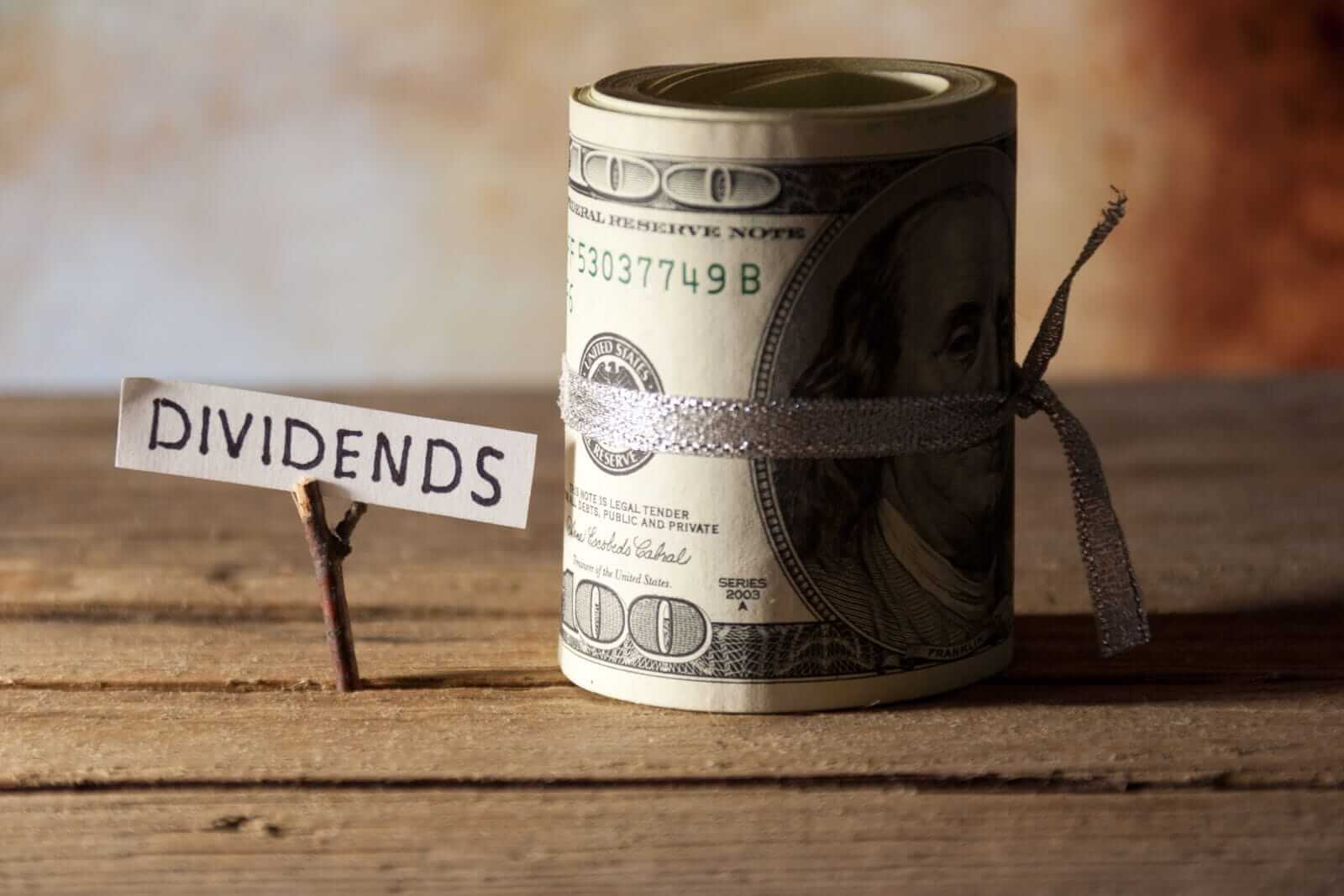Roll of hundred-dollar bills tied with a ribbon next to a small sign labeled 'Dividends'