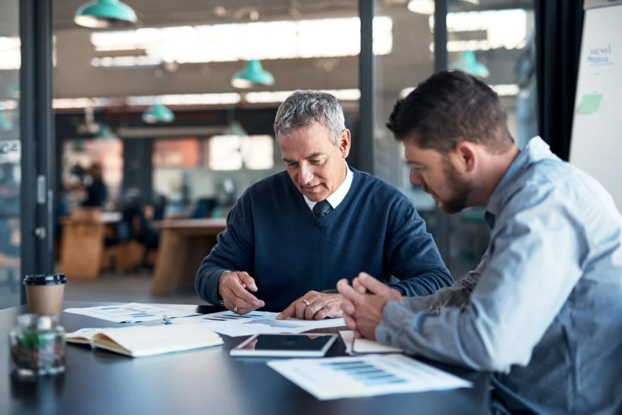 Two businessmen in a modern office reviewing financial documents and charts together at a table