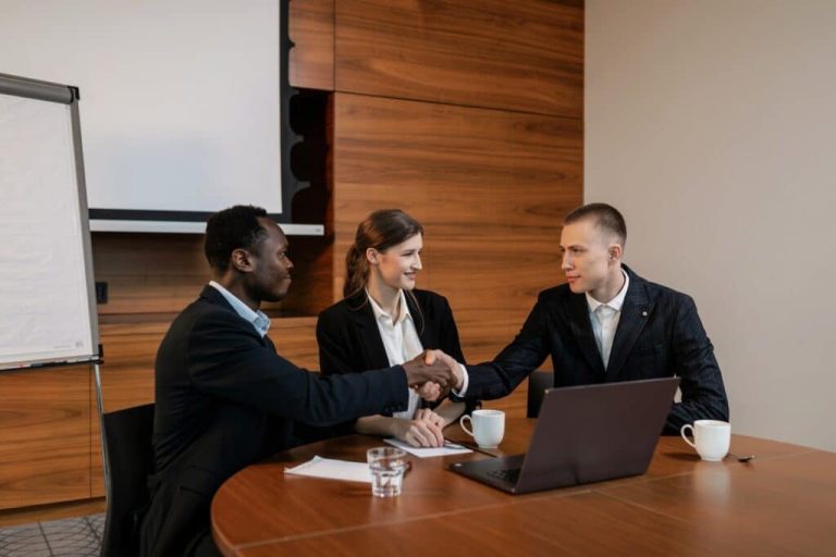 Two businessmen shaking hands at a meeting while a woman looks on and smiles