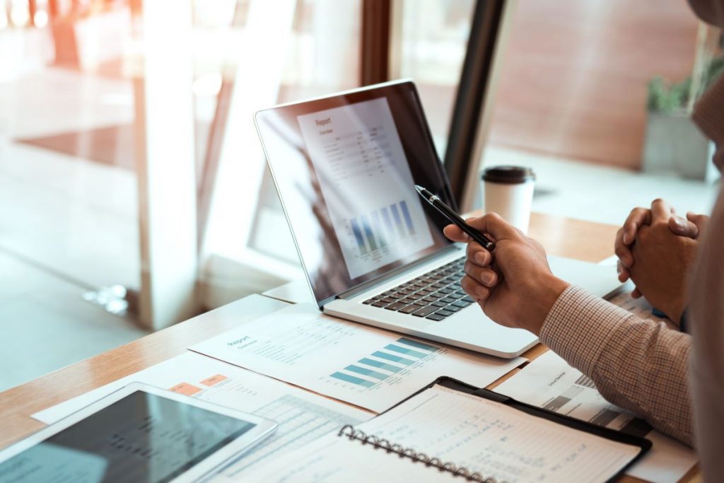 Two professionals reviewing financial charts and data on a laptop at a desk with reports and a tablet.