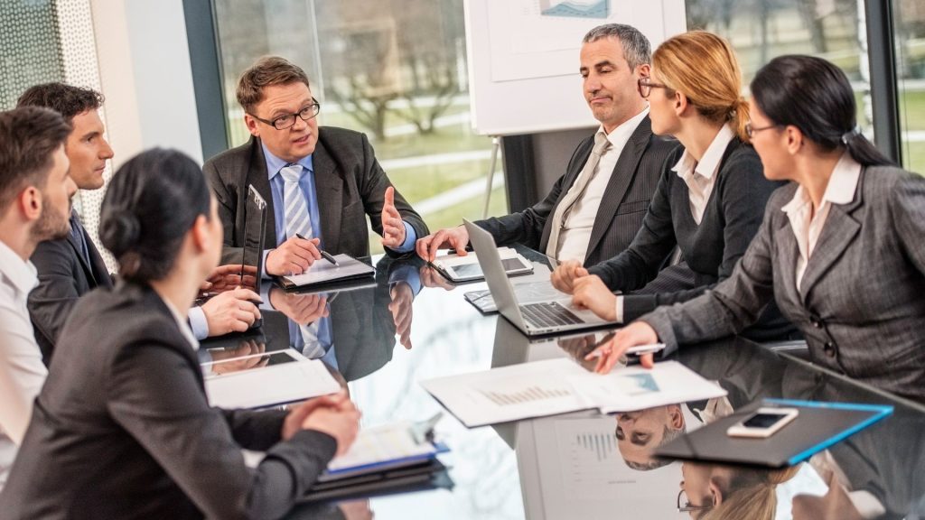 Business professionals in formal attire sitting around a conference table engaged in a serious discussion with documents and laptops
