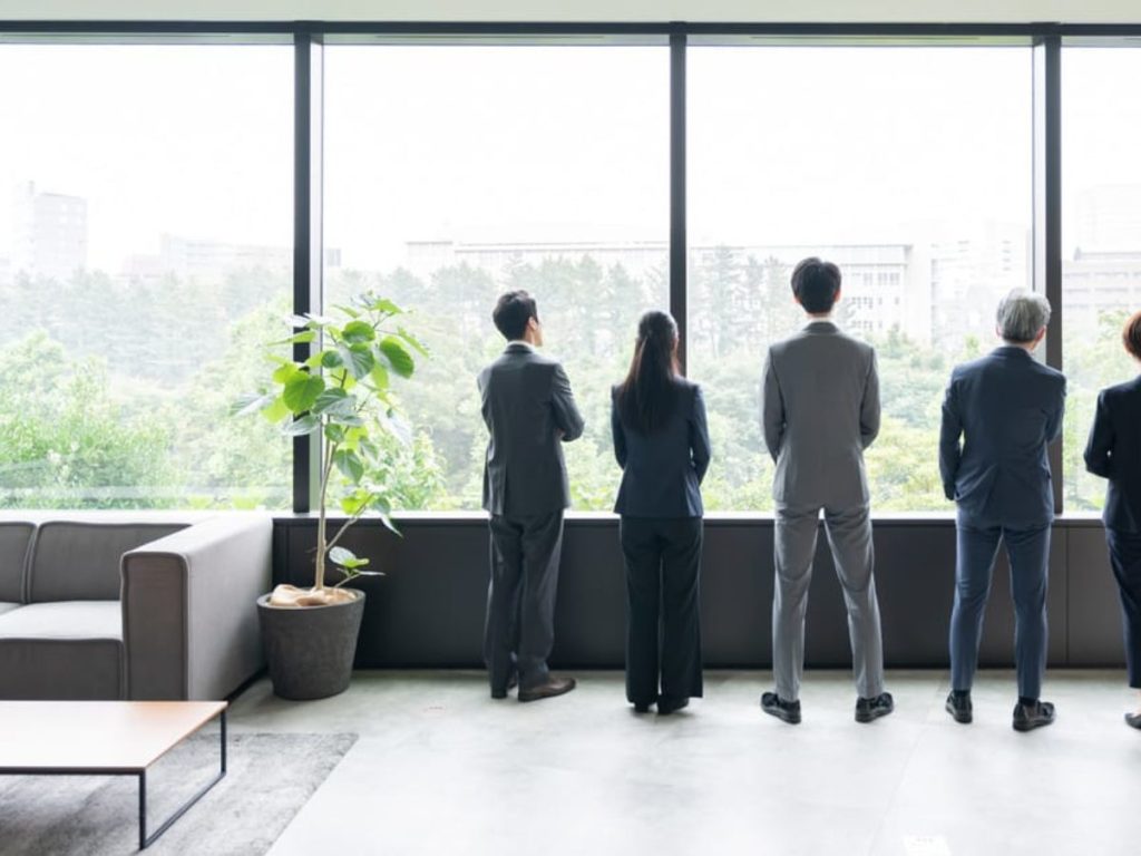 Five business professionals in suits standing and looking out of large office windows with a city and greenery view