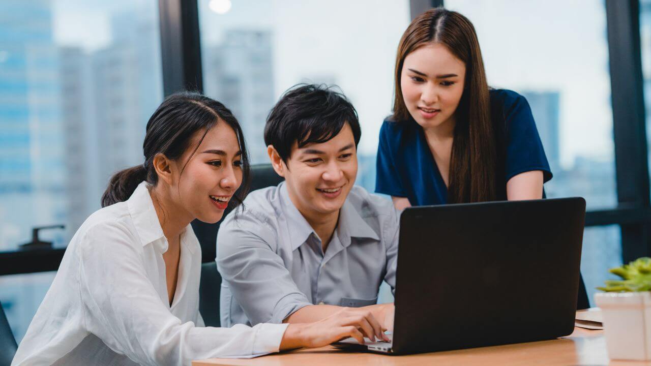 Three young professionals smiling and collaborating while looking at a laptop in a modern office setting