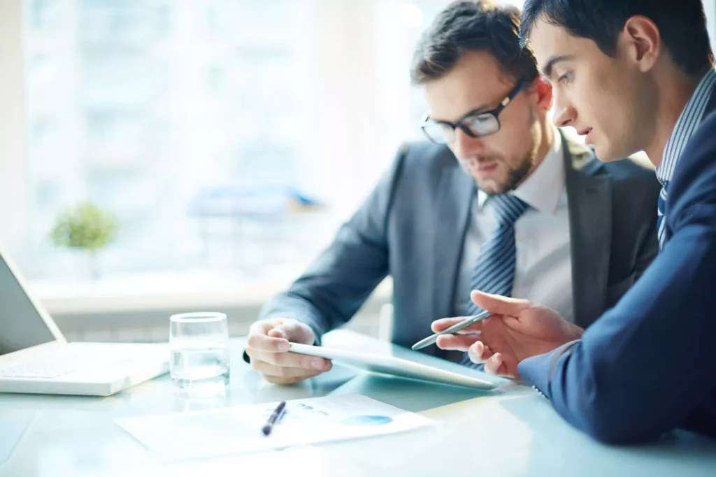 Two businessmen in suits discussing data on a digital tablet during a meeting.
