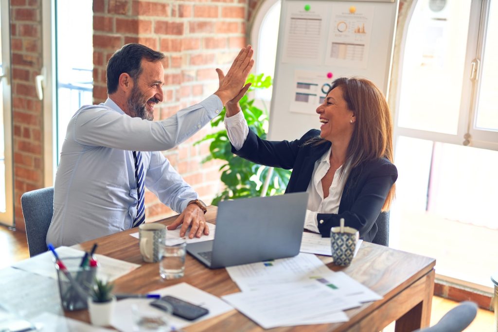 Two cheerful coworkers high-fiving at a desk in a bright office, celebrating a successful achievement
