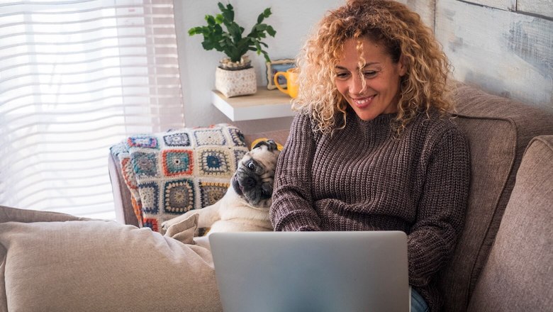 Woman with curly hair relaxing on a couch with her pug, smiling while using a laptop